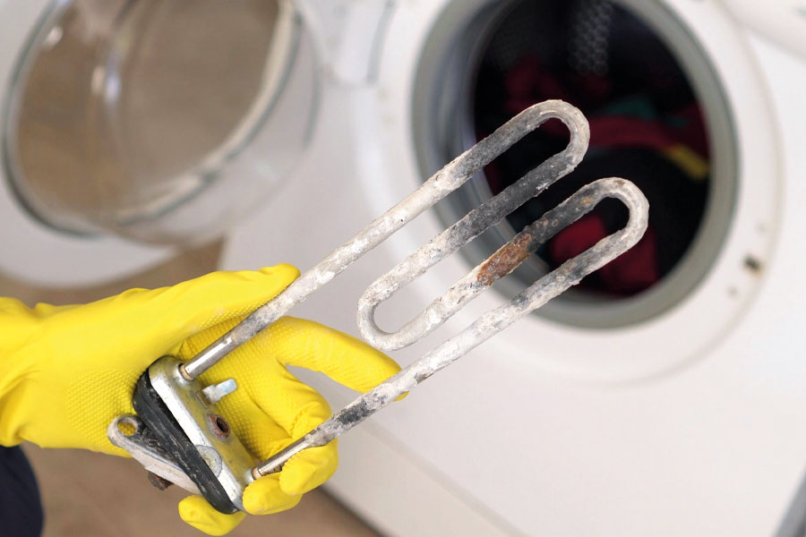 Stark verkalkter Heizstab einer Waschmaschine, ausgebaut und in der Hand gehalten – im Hintergrund die geöffnete Waschmaschine.